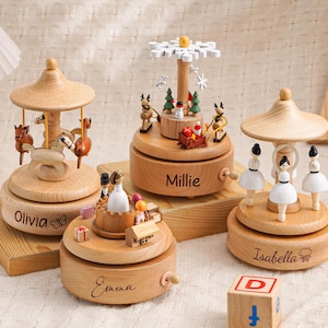 May include: A collection of wooden carousel music boxes featuring different themes, such as horses, a winter scene, and ballerinas. Each box is uniquely designed and engraved with the names "Olivia", "Millie", "Emma", and "Isabella". A wooden block with the letter "D" is also visible.