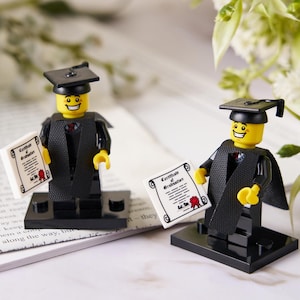 May include: Two Lego figures dressed in graduation attire, including black caps and gowns, stand on black bases. Each figure holds a miniature diploma with a red seal. The figures have yellow heads and are smiling.