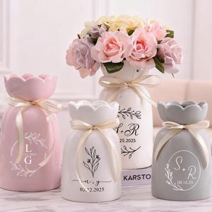 Puede incluir: Cuatro jarrones de cerámica en rosa, blanco y gris, cada uno con un diseño floral único y una cinta. Los jarrones están llenos de flores. Los jarrones tienen texto personalizado con fechas e iniciales.