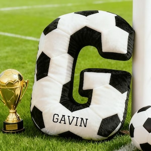 Puede incluir: Un cojín con forma de letra "G" con diseño de balón de fútbol y el nombre "GAVIN" impreso. También se ven un trofeo dorado y un balón de fútbol. La escena se desarrolla en un campo verde.