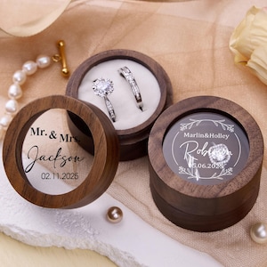 Op de afbeelding: Drie ronde houten ringendoosjes met doorzichtige deksels. Twee dozen bevatten diamanten ringen. Eén doos heeft de tekst "Mr. & Mrs. Jackson 02.11.2025". Een andere doos heeft de tekst "Marlin & Holley Robin 05.06.2025".