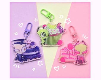 May include: Three acrylic keychains featuring cartoon characters. One keychain shows a character with a purple dessert, another with a cupcake, and the last with a pink cake. Each keychain has a different colored clasp: purple, green, and pink.