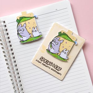 Puede incluir: Dos marcadores magn&eacute;ticos con una ilustraci&oacute;n de dibujos animados de dos personajes debajo de una hoja. Los marcadores son de color lila claro, blanco y verde, con el texto "SPRING LEAF ICONIC MAGNETIC BOOKMARKS" en uno. Est&aacute;n colocados en un cuaderno de espiral.
