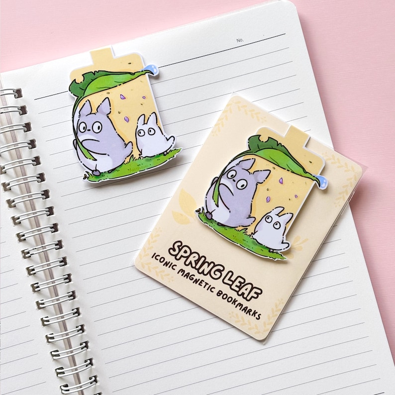 Puede incluir: Dos marcadores magn&eacute;ticos con una ilustraci&oacute;n de dibujos animados de dos personajes debajo de una hoja. Los marcadores son de color lila claro, blanco y verde, con el texto "SPRING LEAF ICONIC MAGNETIC BOOKMARKS" en uno. Est&aacute;n colocados en un cuaderno de espiral.