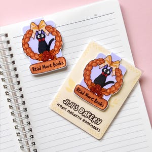 Puede incluir: Dos marcadores magn&eacute;ticos con un gato negro con pajarita roja dentro de una corona con un lazo amarillo. Los marcadores tienen el texto "Read More Books" y "JIJI'S BAKERY ICONIC MAGNETIC BOOKMARKS". Los marcadores est&aacute;n en un cuaderno.