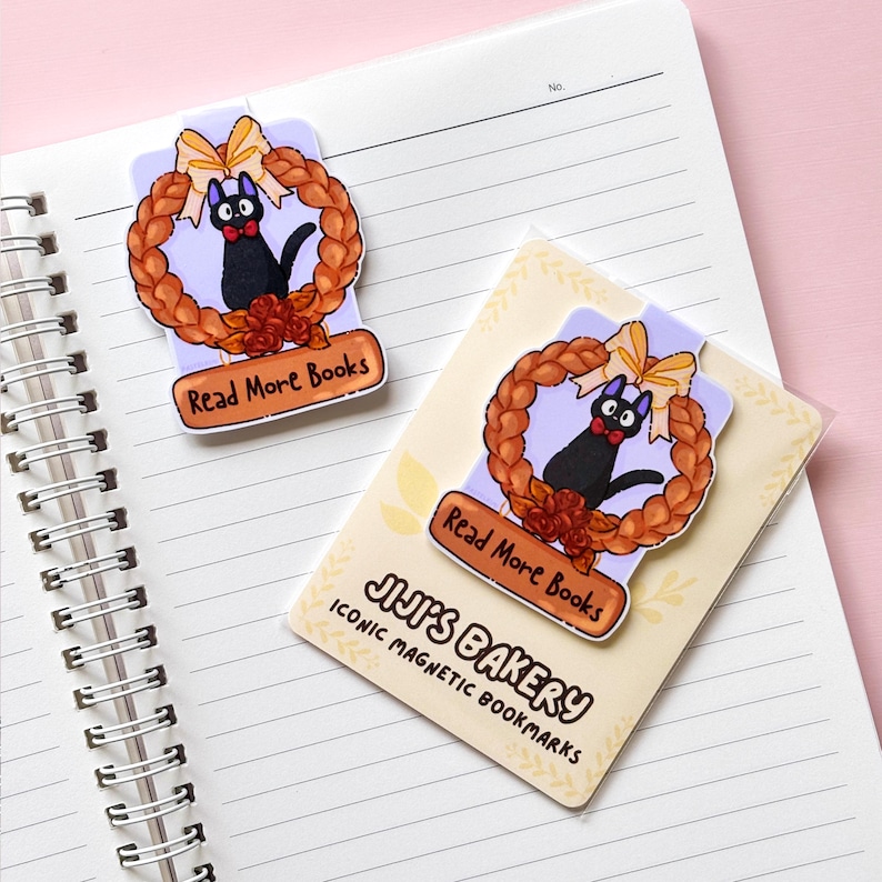 Puede incluir: Dos marcadores magn&eacute;ticos con un gato negro con pajarita roja dentro de una corona con un lazo amarillo. Los marcadores tienen el texto "Read More Books" y "JIJI'S BAKERY ICONIC MAGNETIC BOOKMARKS". Los marcadores est&aacute;n en un cuaderno.