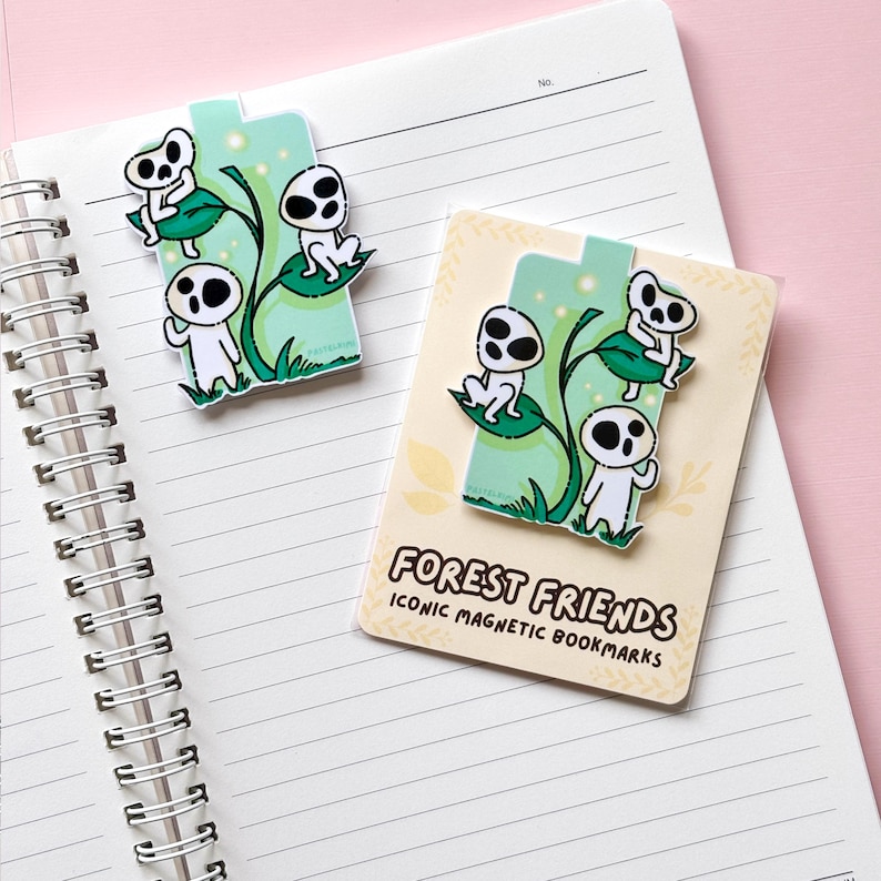 Puede incluir: Dos marcap&aacute;ginas magn&eacute;ticos "Forest Friends" en un cuaderno. Cada marcap&aacute;ginas presenta una ilustraci&oacute;n de dibujos animados de tres figuras blancas con ojos negros y un dise&ntilde;o de hojas. El texto "FOREST FRIENDS ICONIC MAGNETIC BOOKMARKS" est&aacute; en un marcap&aacute;ginas.
