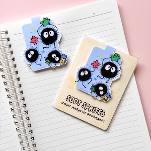 Puede incluir: Marcadores magn&eacute;ticos azul claro con Soot Sprites, con cuerpos negros y redondos y grandes ojos blancos. Los marcadores tienen detalles de estrellas y flores. El texto "SOOT SPRITES ICONIC MAGNETIC BOOKMARKS" es visible.
