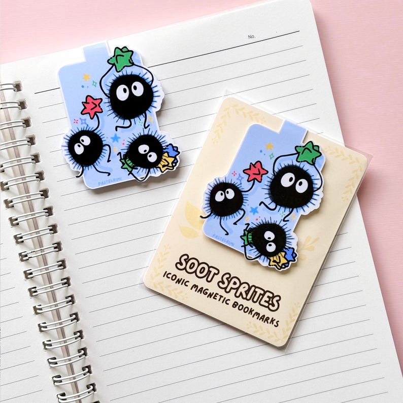 Puede incluir: Marcadores magn&eacute;ticos azul claro con Soot Sprites, con cuerpos negros y redondos y grandes ojos blancos. Los marcadores tienen detalles de estrellas y flores. El texto "SOOT SPRITES ICONIC MAGNETIC BOOKMARKS" es visible.