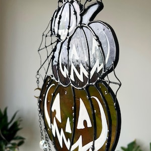 May include: A stained glass Halloween decoration featuring two stacked pumpkins with carved faces. The pumpkins are in shades of green and brown with white accents and are connected by a black chain. A spiderweb design is visible.