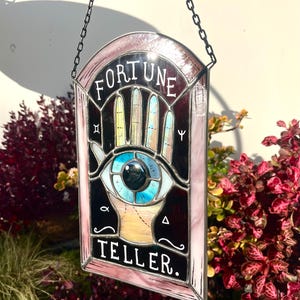 Könnte beinhalten: Buntglasscheibe mit dem Thema Wahrsager. Das Design zeigt ein Auge, eine Hand und die Worte "FORTUNE TELLER". Die Scheibe ist rosa umrandet und hängt an einer schwarzen Kette. Das Auge hat eine blaue Iris und eine schwarze Pupille.