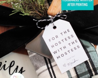 May include: A gift set with a white tag that reads "FOR THE HOSTESS WITH THE MOSTESS". The tag is tied with twine to a black ribbon and a wooden handle. The gift is wrapped with a plaid cloth and greenery.