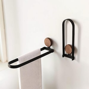 May include: A black metal towel rack with a wooden accent, holding a white towel. A matching wall hook with a wooden accent is next to the towel rack. The towel rack is approximately 12 inches wide.