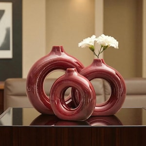 Può includere: Tre vasi in ceramica rossa di diverse dimensioni, ciascuno con un'apertura circolare al centro. Un vaso contiene fiori bianchi. I vasi hanno una finitura lucida e sono disposti su una superficie scura.