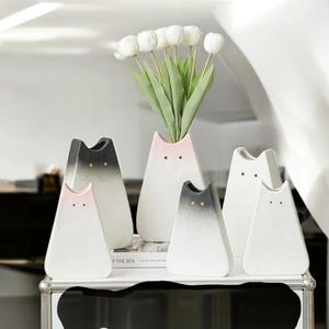 Könnte beinhalten: Fünf Keramikvasen in Katzenform in verschiedenen Farben, darunter Weiß, Schwarz und Rosa. Die Vasen sind auf einer schwarzen Oberfläche mit einem Buch davor angeordnet. Ein Strauß weißer Blumen steht in der mittleren Vase.