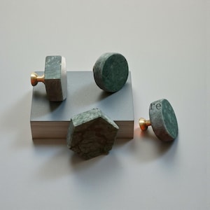 May include: Four decorative cabinet knobs. The knobs are made of green marble with brass accents. One knob is a cube shape, one is a cylinder, one is a geometric shape, and one is a semi-circle. The knobs are on a gray surface.