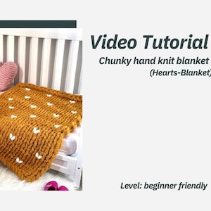 May include: A mustard yellow chunky knit blanket with white heart-shaped details. The blanket is on a white cot with two pink and white heart-shaped cushions. Video Tutorial: Chunky hand knit blanket (Hearts-Blanket) Level: beginner friendly