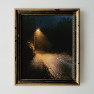 May include: A framed painting depicting a dark, nighttime road illuminated by a single glowing street lamp. The scene features deep shadows, silhouettes of trees, and a warm, golden light cast onto the pavement, all encased in a dark wood frame.