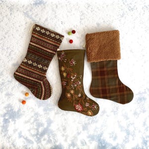 May include: Three Christmas stockings in shades of brown and green rest on a snowy surface. One stocking has a patterned design, another features floral embroidery, and the third has a plaid pattern with a faux fur cuff.