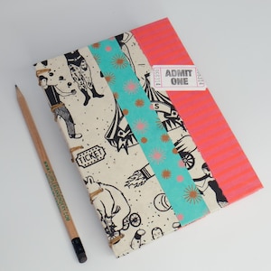 Könnte beinhalten: Ein handgefertigtes Notizbuch mit Zirkusmotiv. Der Einband hat einen cremefarbenen Hintergrund mit schwarz-weißen Zirkusillustrationen. Ein türkisfarbener Streifen mit rosa floralen Akzenten und ein rosa Ticket mit der Aufschrift "ADMIT ONE" befinden sich auf dem Einband. Ein Bleistift liegt daneben.