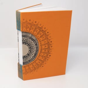May include: An orange notebook with a black and white mandala design on the cover. The notebook is closed and the spine is visible.