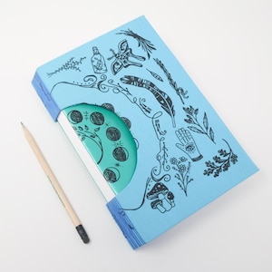 Può includere: Un diario azzurro con un ritaglio che rivela una copertina circolare color ottanio con fasi lunari. La copertina blu è decorata con disegni a linee nere di una falena, una piuma, una mano con un occhio, funghi e piante. Una matita di legno è posizionata accanto.