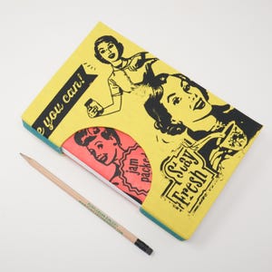 May include: A yellow notebook with retro illustrations and the text "Stay Fresh" and "e you can!". A pencil with the text "PURPLEBEAN BINDERY" lies next to the notebook. The notebook has a red and white interior with the text "jam packed".