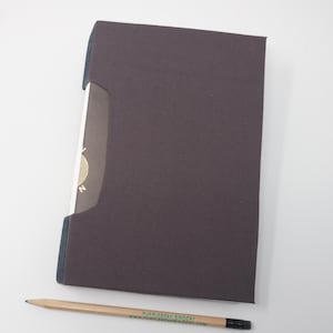 Puede incluir: Un libro de tapa dura de color marrón oscuro con un diseño dorado en el lomo. El libro está cerrado y acostado sobre una superficie blanca. Un lápiz está acostado al lado del libro. El lápiz tiene el texto "Purple Bean Bindery" y una dirección de sitio web impresa.