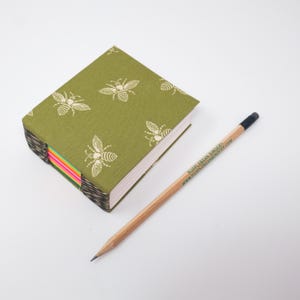May include: A green notebook with a bee pattern and a pencil. The notebook has a green cover with a repeating pattern of cream-colored bees. The pencil is wooden with a black eraser and the text "PURPLEBEAN BINDER" and website address.