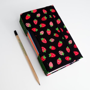 May include: A black notebook with a strawberry pattern, featuring red and pink strawberries with green stems and faces. A wooden pencil with black lead and text is next to the notebook. The notebook has a colorful edge and an elastic band.