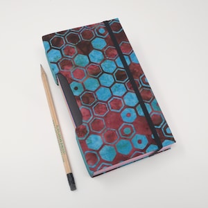 Puede incluir: Un cuaderno con una cubierta con estampado de panal de abeja azul y granate, una banda elástica negra y un lápiz. El lápiz tiene el texto "PURPLEBEAN BINDERY". El cuaderno está abierto para revelar un bolsillo negro.