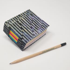 Puede incluir: Un pequeño libro cuadrado con una cubierta de patrón blanco y negro y bordes de página expuestos en colores arcoíris. Junto al libro hay un lápiz de madera clara con goma negra, que presenta el texto impreso PURPLEBEAN BINDERY.