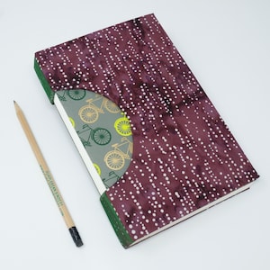 May include: A hardbound notebook with a burgundy cover featuring a white dot pattern. The spine is green, and the inside cover has a bicycle design in green and gold. A pencil rests beside the notebook.