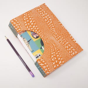 May include: A rectangular notebook with an orange cover featuring a white pattern of circles and crosses. The cover has a large semi-circular cutout revealing a multi-colored graphic print on the inner page. A purple pencil lies beside it.