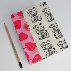 Könnte beinhalten: Ein handgefertigtes Notizbuch mit Erdbeer- und Glückskatzen-Design. Das Notizbuch hat einen rosa Rücken mit roten Erdbeeren und einen cremefarbenen Einband mit schwarz-weißen Glückskatzen-Illustrationen. Ein Bleistift mit Text liegt neben dem Notizbuch.