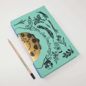 May include: A teal journal with a gold-colored edge and a pencil. The journal cover features black illustrations of plants, feathers, and a hand with an eye. The pencil has the text "PURPLEBEAN BINDERY" printed on it.