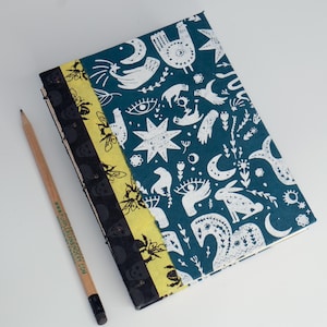 May include: A handmade journal with a teal cover featuring white celestial and animal illustrations. The spine has a black and yellow patterned design. A wooden pencil with the text "www.purplebeanbindery.com" rests beside the journal.