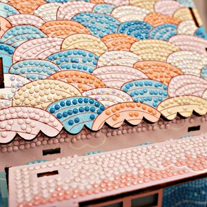 May include: A wooden craft kit with a pink and white roof covered in colorful round beads. The roof is shaped like a series of overlapping waves.