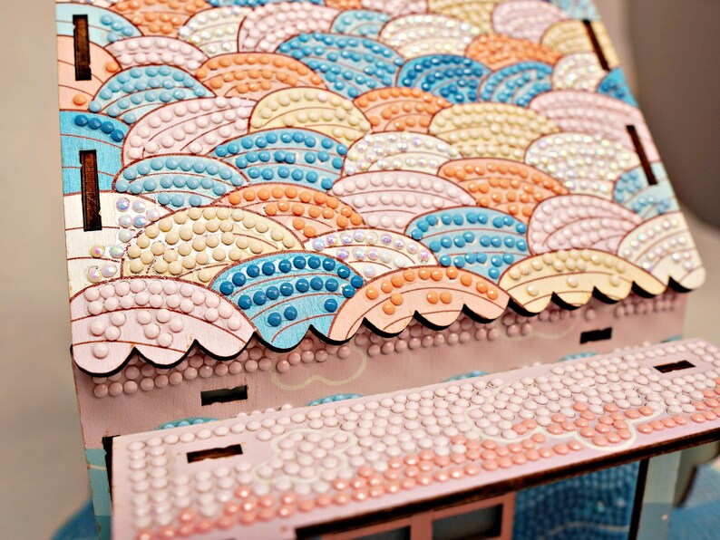 May include: A wooden craft kit with a pink and white roof covered in colorful round beads. The roof is shaped like a series of overlapping waves.