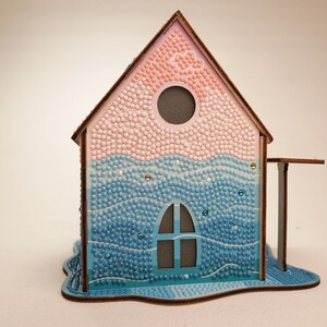 May include: A wooden house model with a pink and blue mosaic design. The house has a window and a round opening in the roof. The base of the house is shaped like a wave.