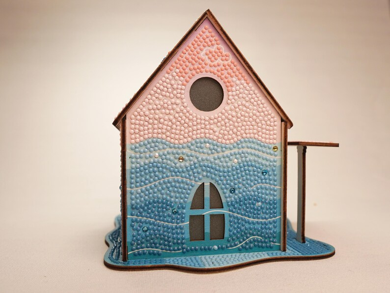 May include: A wooden house model with a pink and blue mosaic design. The house has a window and a round opening in the roof. The base of the house is shaped like a wave.