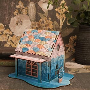 May include: A small wooden house model with a blue, pink, and orange mosaic roof. The house has a porch with a window and a small tree in front.