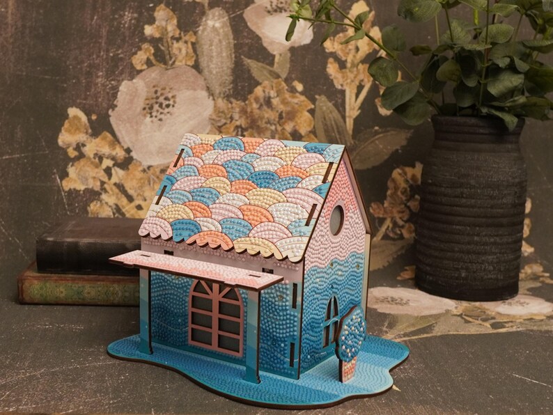 May include: A small wooden house model with a blue, pink, and orange mosaic roof. The house has a porch with a window and a small tree in front.