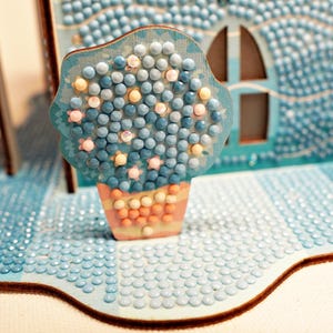 May include: A blue and white diamond painting of a house with a blue flower pot and a blue flower in it.