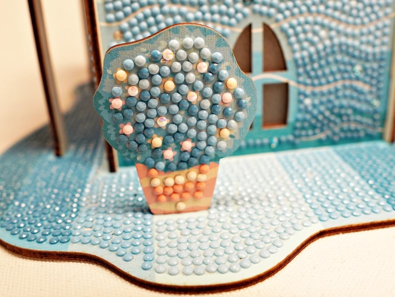May include: A blue and white diamond painting of a house with a blue flower pot and a blue flower in it.