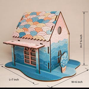 May include: A wooden 3D model of a house with a blue and pink paint scheme. The roof is decorated with a pattern of overlapping scales. The house is 7 1/4 inches tall, 7 inches long, and 6 inches wide.