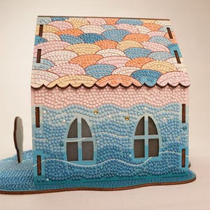 May include: A wooden house shaped 3D diamond painting kit with a pink and blue color scheme. The roof is decorated with a pattern of overlapping pastel-colored circles. The house has two windows and a base that looks like waves.