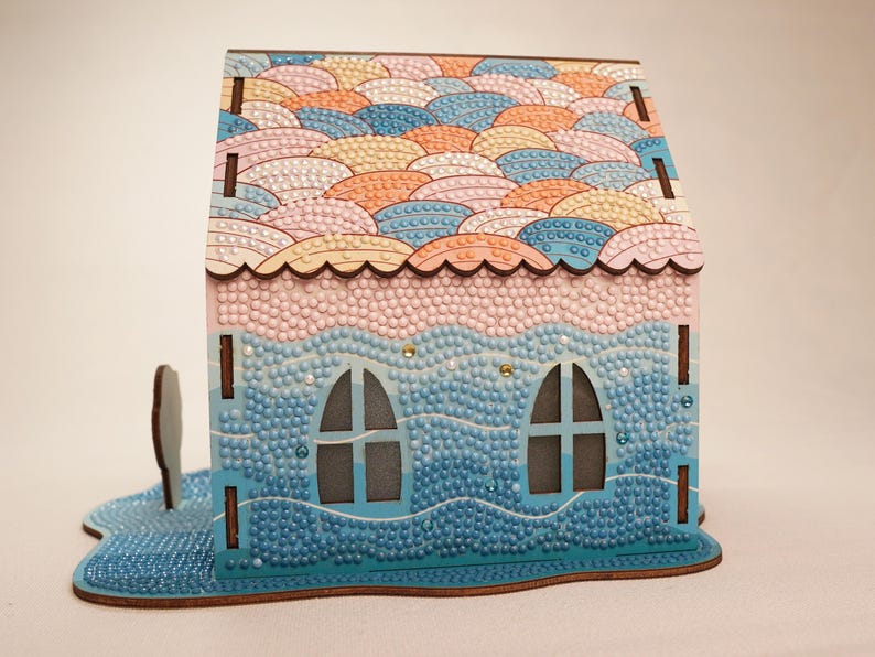 May include: A wooden house shaped 3D diamond painting kit with a pink and blue color scheme. The roof is decorated with a pattern of overlapping pastel-colored circles. The house has two windows and a base that looks like waves.