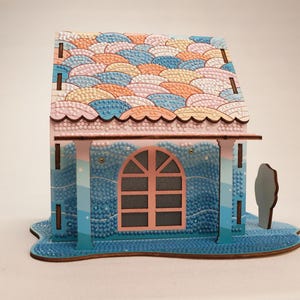 May include: A small wooden house with a pink roof and blue walls. The roof is decorated with a pattern of colorful waves. The house has a single window with a pink frame and a blue door. The house is sitting on a blue base.