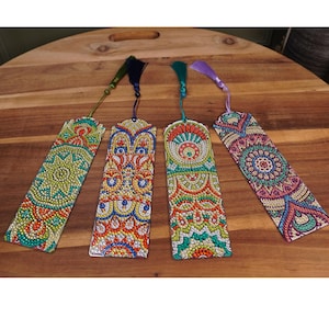 May include: Four colorful, patterned bookmarks with tassels. Each bookmark features a unique design with intricate details in shades of green, orange, blue, and purple. The tassels are in matching colors, adding a decorative touch to these reading accessories.
